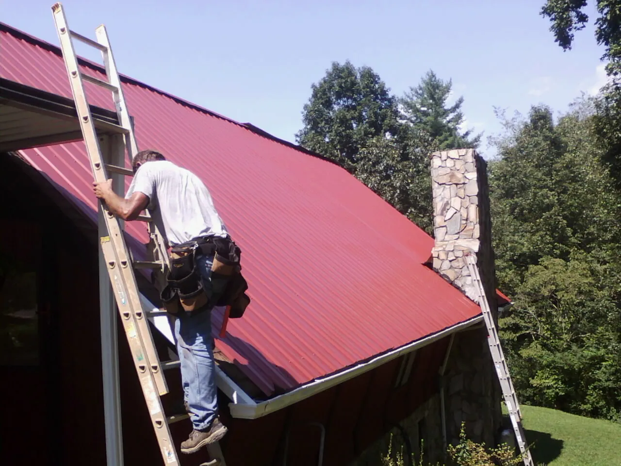 Roofer on a ladder beside a red metal roof and stone chimney — Soffit & Fascia Repair by Roofing Brothers in Southwick